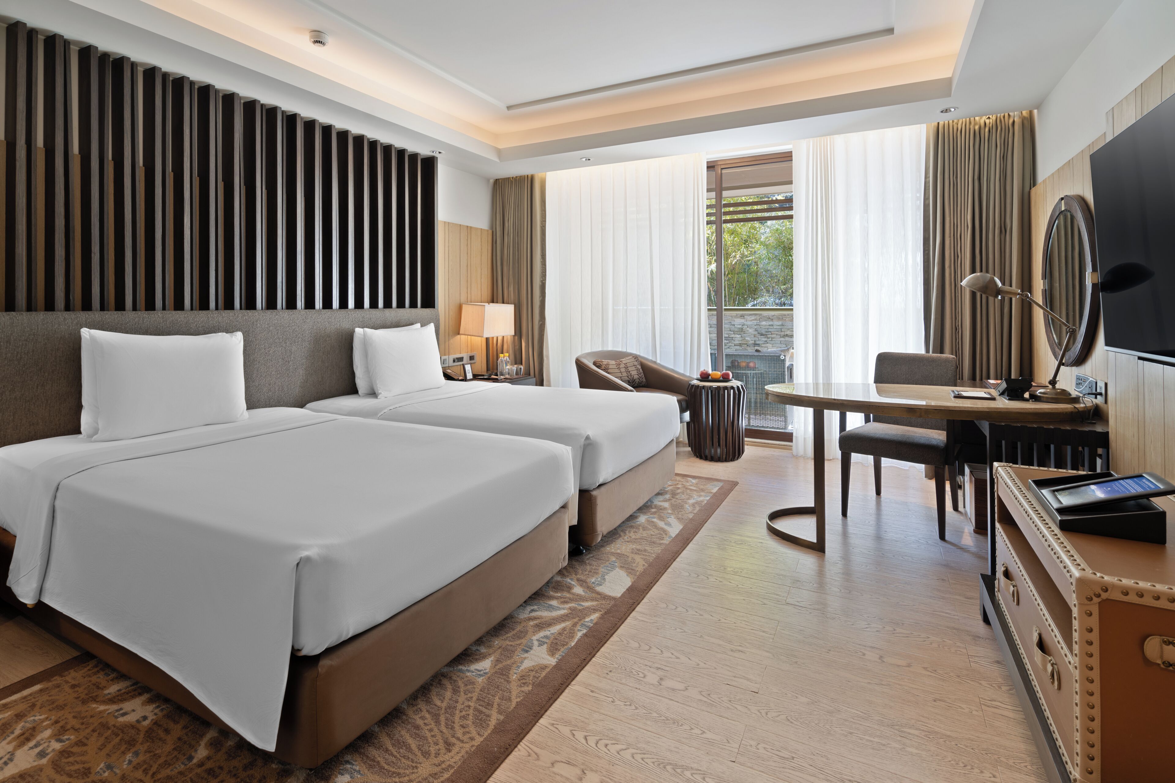 A hotel room with two beds, a desk, a chair, a TV, and a large window with curtains.