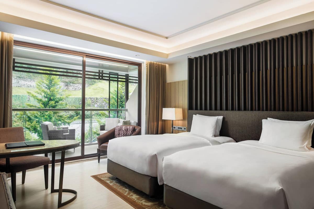 Une chambre d’hôtel avec deux lits, un bureau et une vue sur la verdure grâce à de grandes fenêtres.