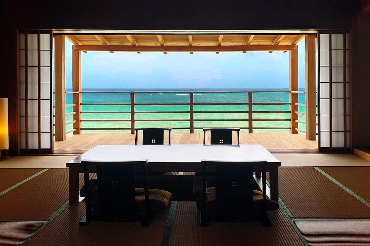 A room with tatami mats, a wooden table, and chairs, with a view of the ocean through a large window.