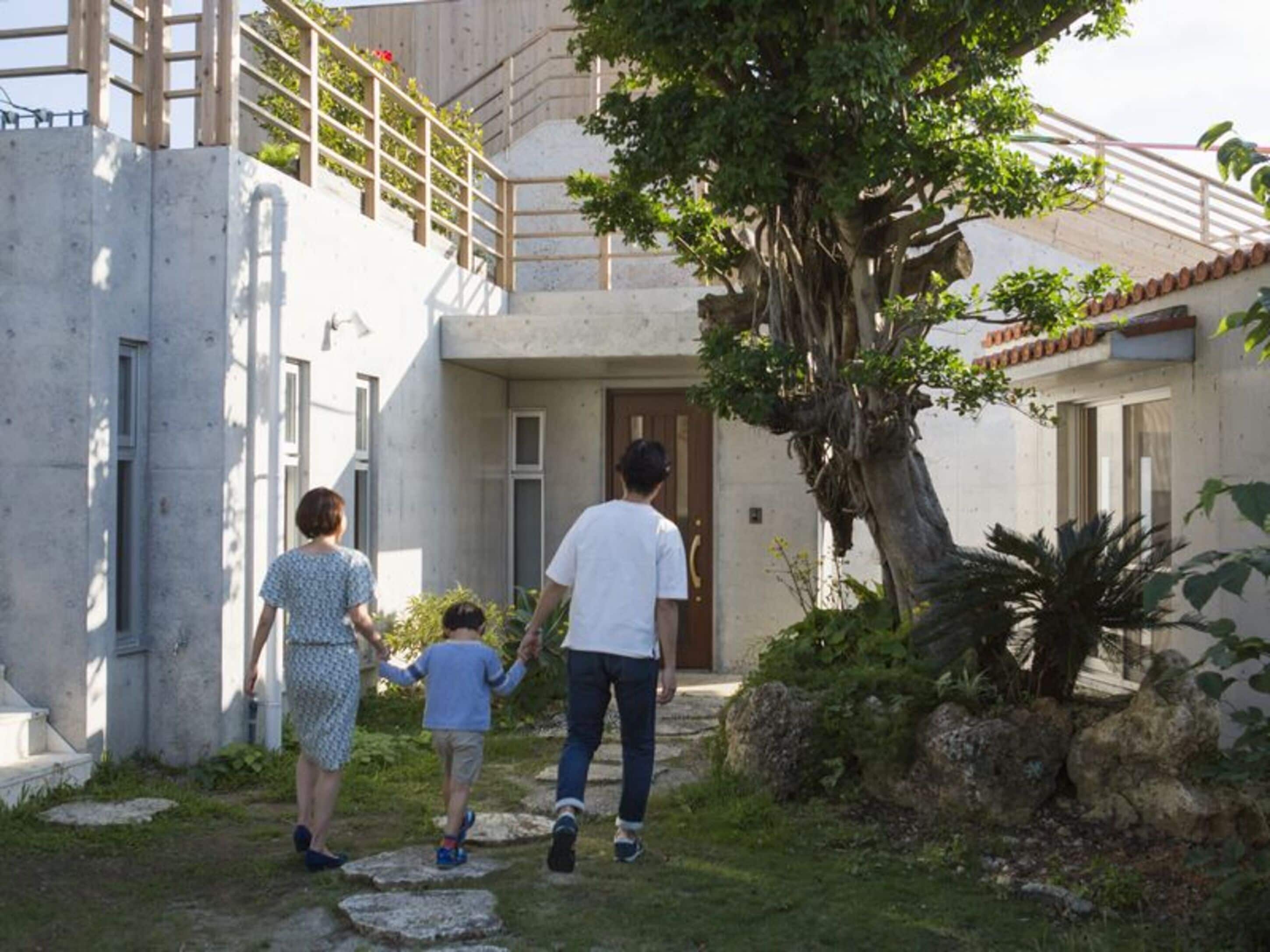 A family walking towards a house with a stone pathway, a large tree, and a wooden fence.