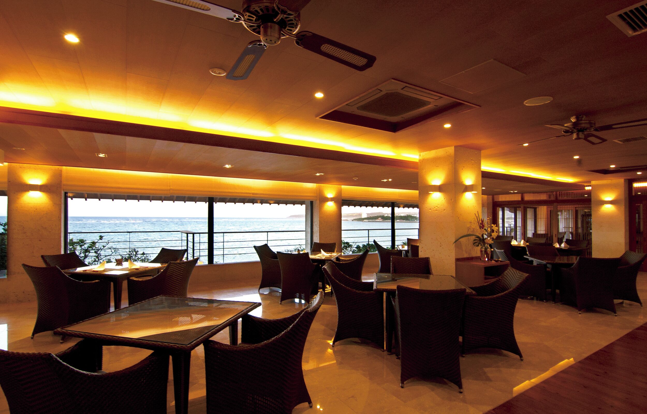 A restaurant with tables and chairs, large windows with a view of the sea, and ceiling fans.