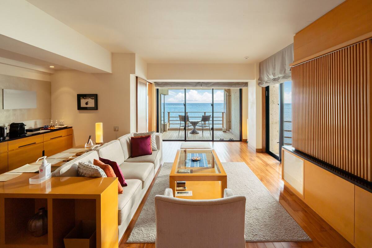 A spacious living room with a view of the sea, a dining area with a table and chairs, and a kitchen area with a coffee machine.