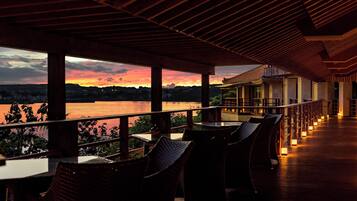 An outdoor dining area with tables and chairs, overlooking a body of water and a sunset.