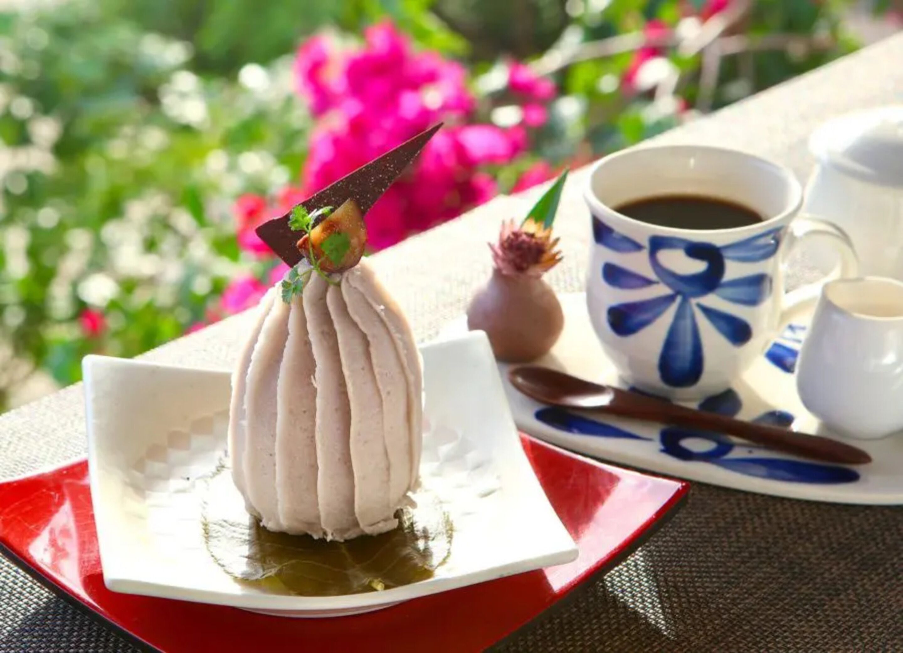 Un postre sobre un plato blanco con base roja, una taza de café y un pequeño florero con una flor en una mesa al aire libre.