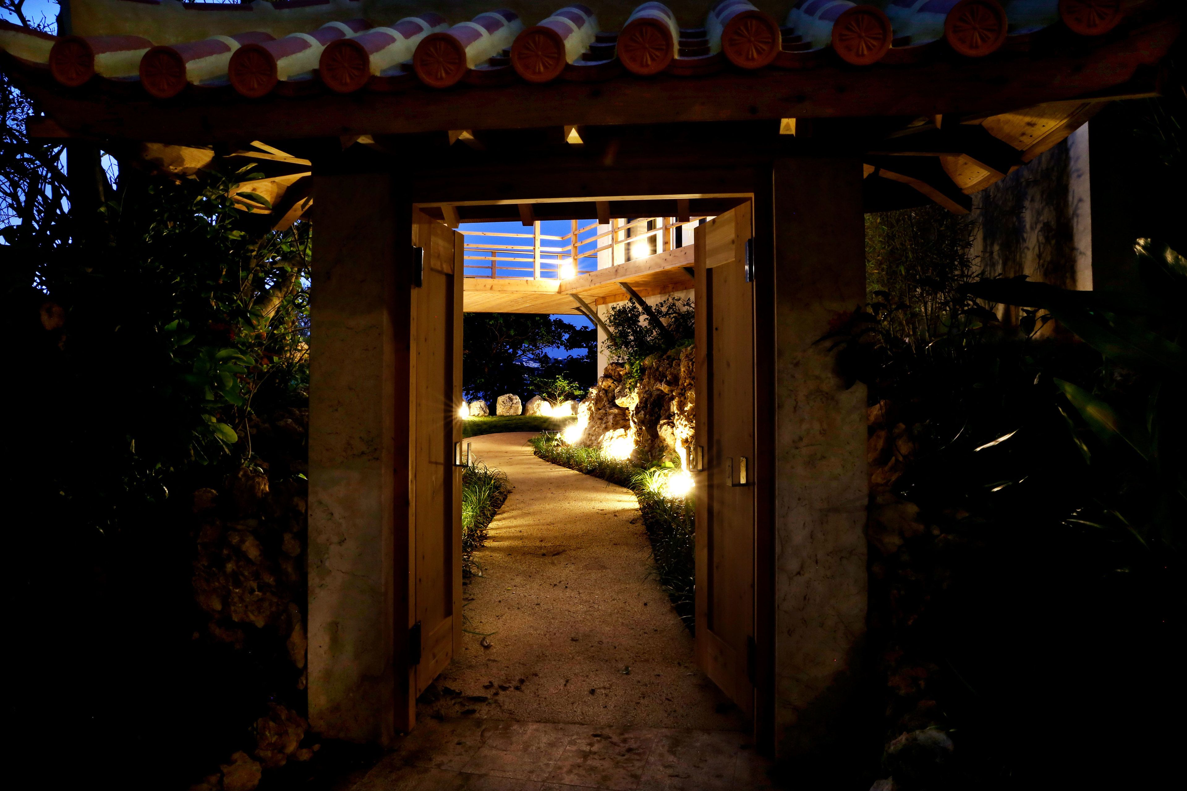 A pathway leading to a wooden structure with a tiled roof, surrounded by greenery and illuminated by ambient lighting.
