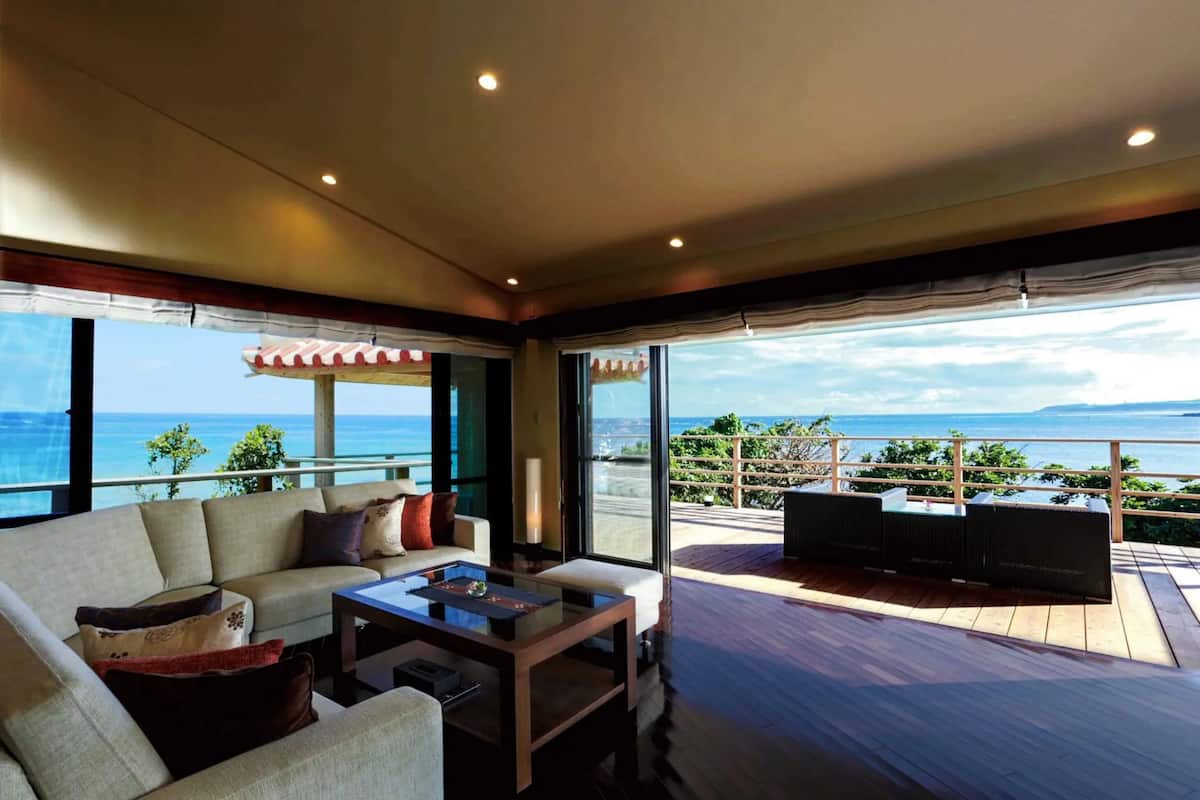 A modern living room with a sofa, coffee table, and a TV. It features large windows with a view of the ocean and a balcony with outdoor seating.