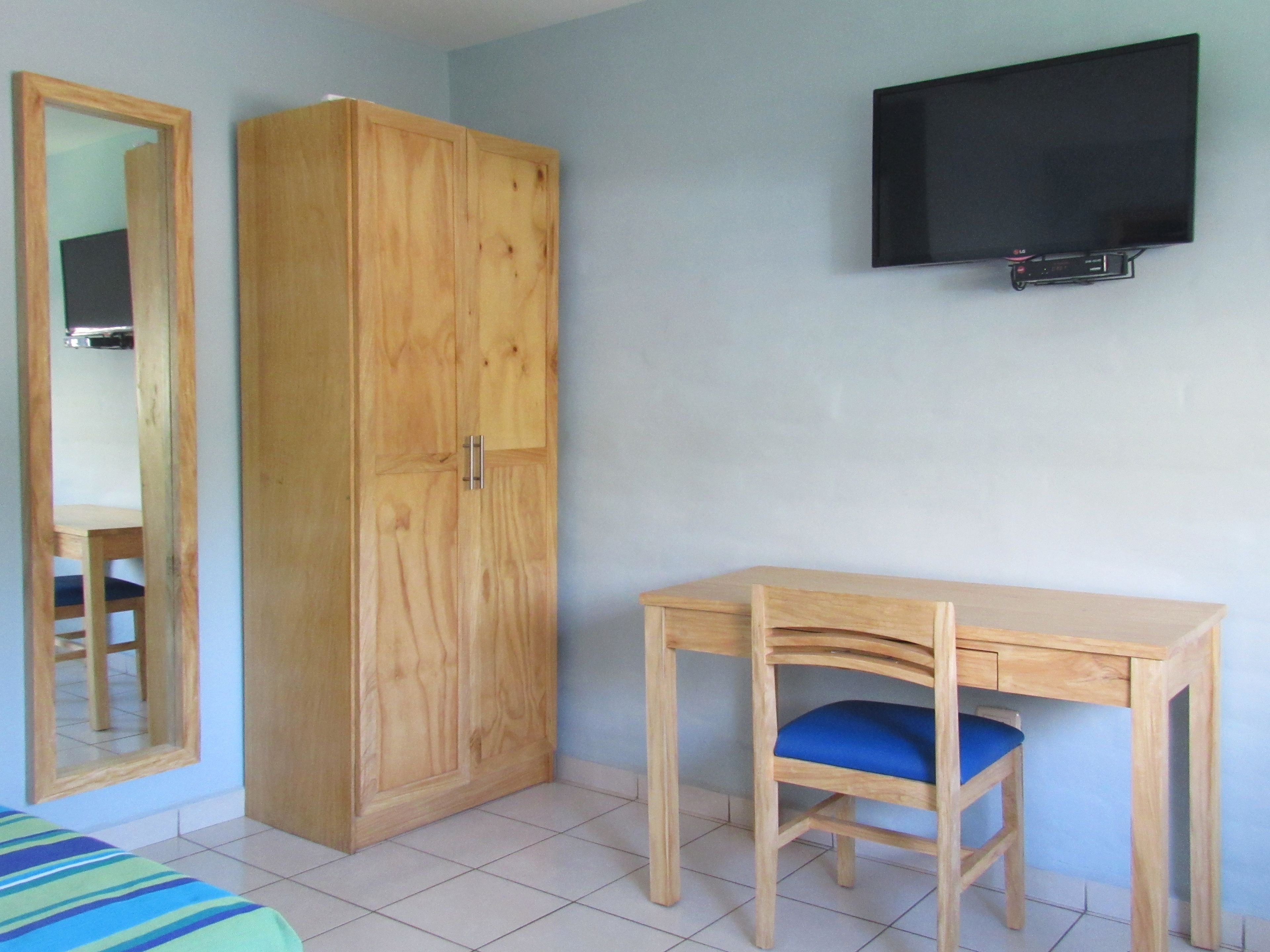 Quarto com guarda-roupa de madeira, escrivaninha com cadeira azul, cama com colcha listrada e televisão fixada na parede.