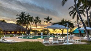 A poolside area with lounge chairs and umbrellas, surrounded by palm trees and a thatched-roof building. - Atami Escape Resort (Tamanique)