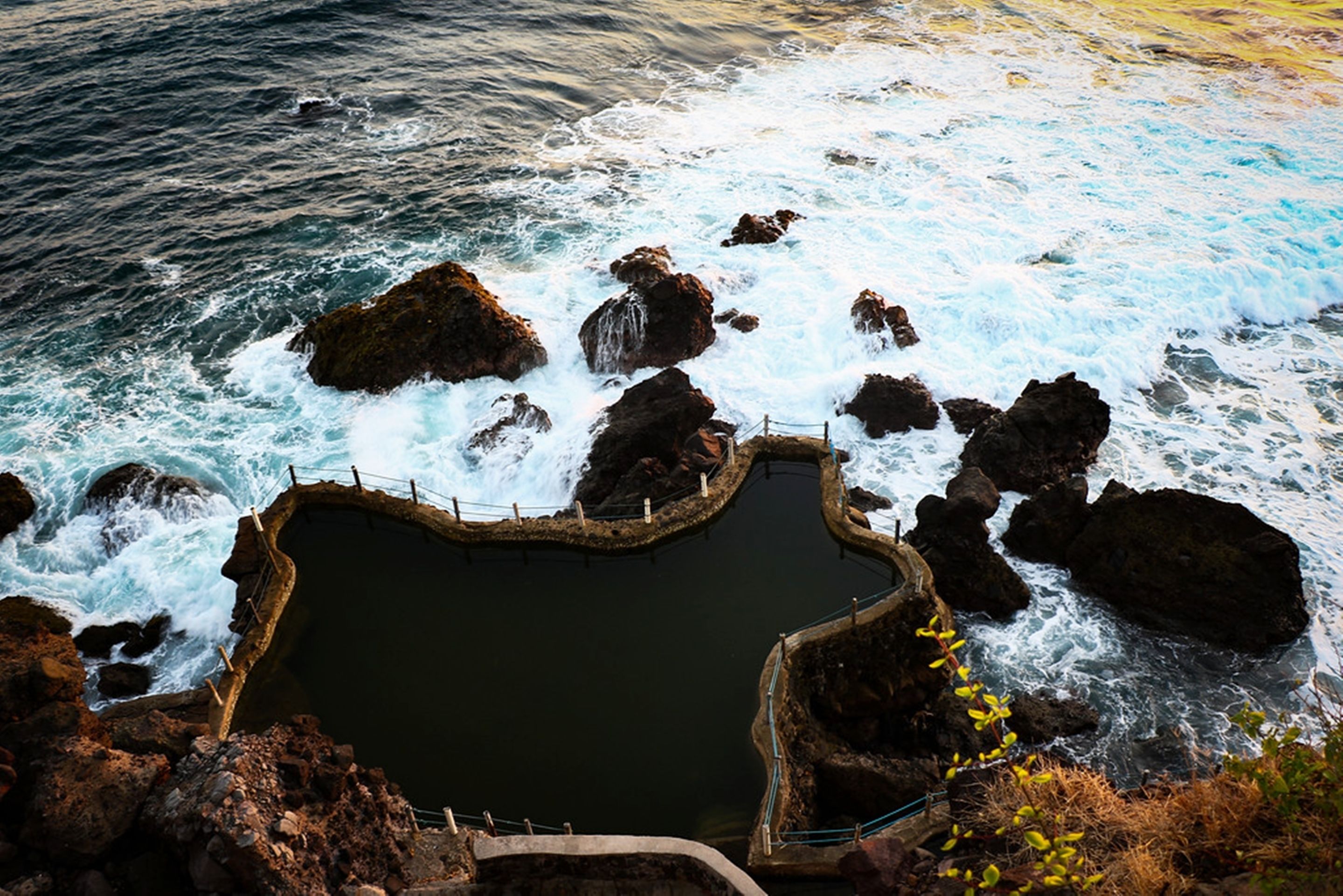A coastal rocky pool with waves crashing against it, surrounded by cliffs and a railing.