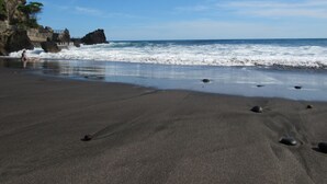 A beach with dark sand, waves crashing onto the shore, and a rocky cliff with vegetation. - Atami Escape Resort (Tamanique)