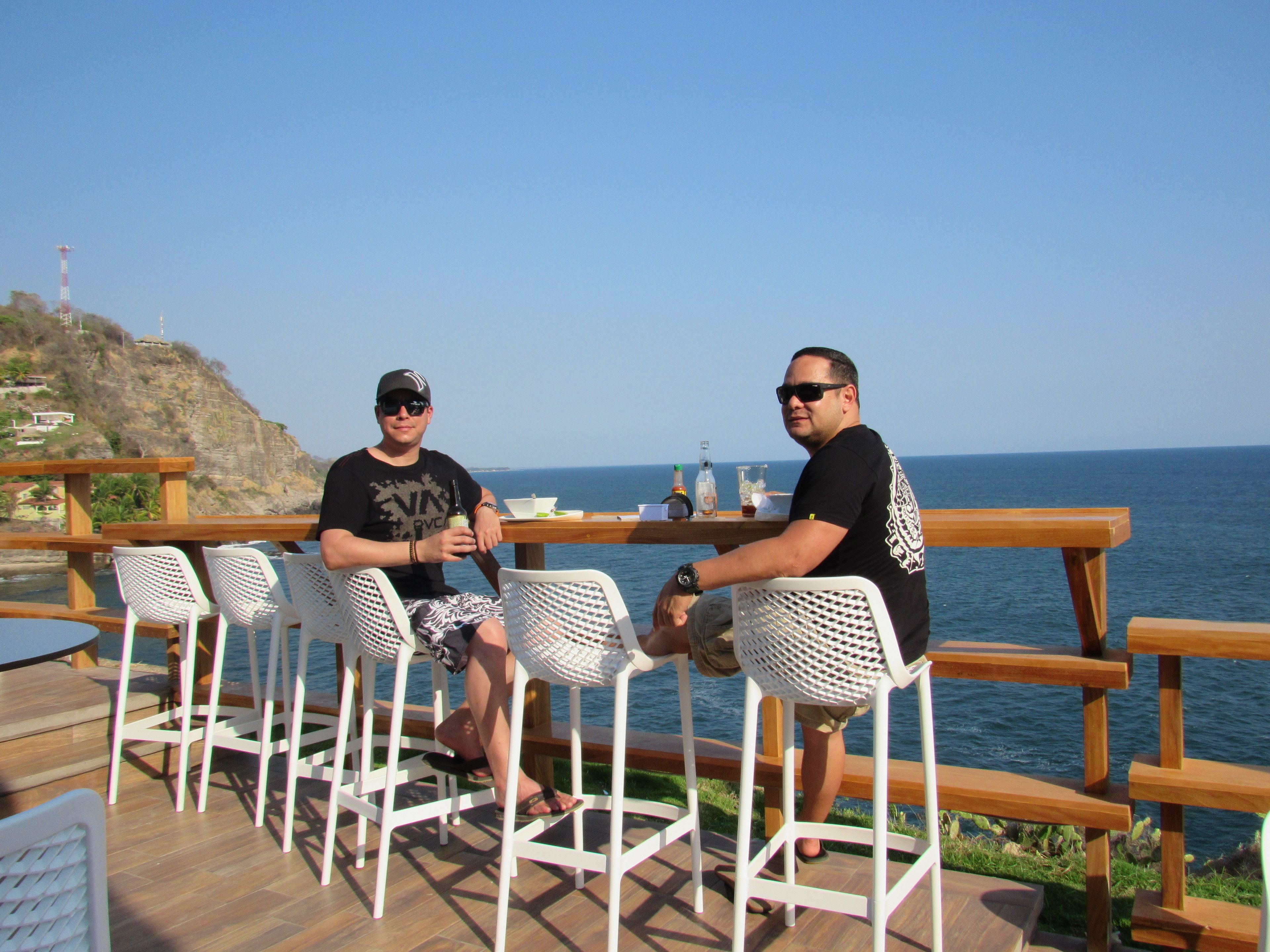 Two men seated at a wooden table with white chairs, overlooking a body of water and a rocky landscape.