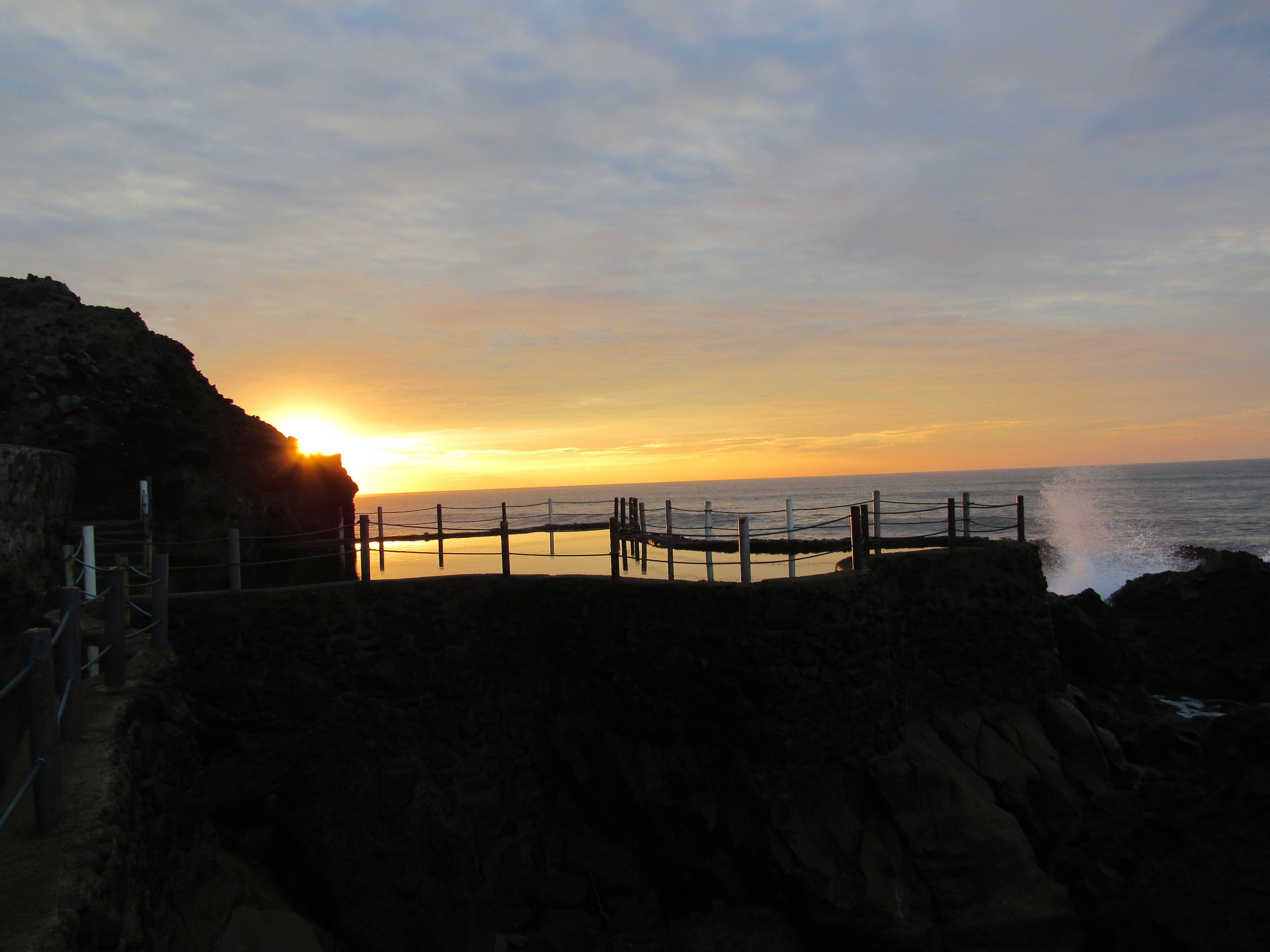A coastal sunset view with a rocky shoreline and a wooden fence.