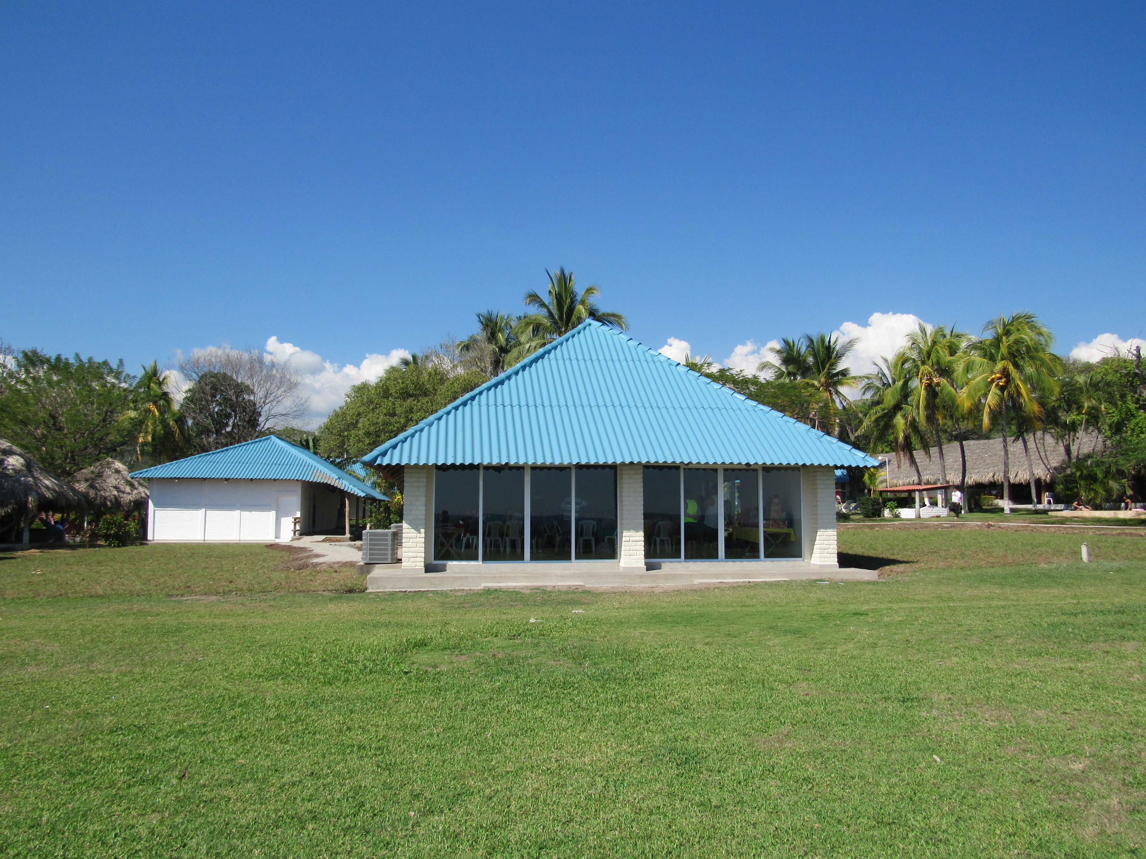 A building with a blue roof and large windows, surrounded by a grassy area and palm trees.