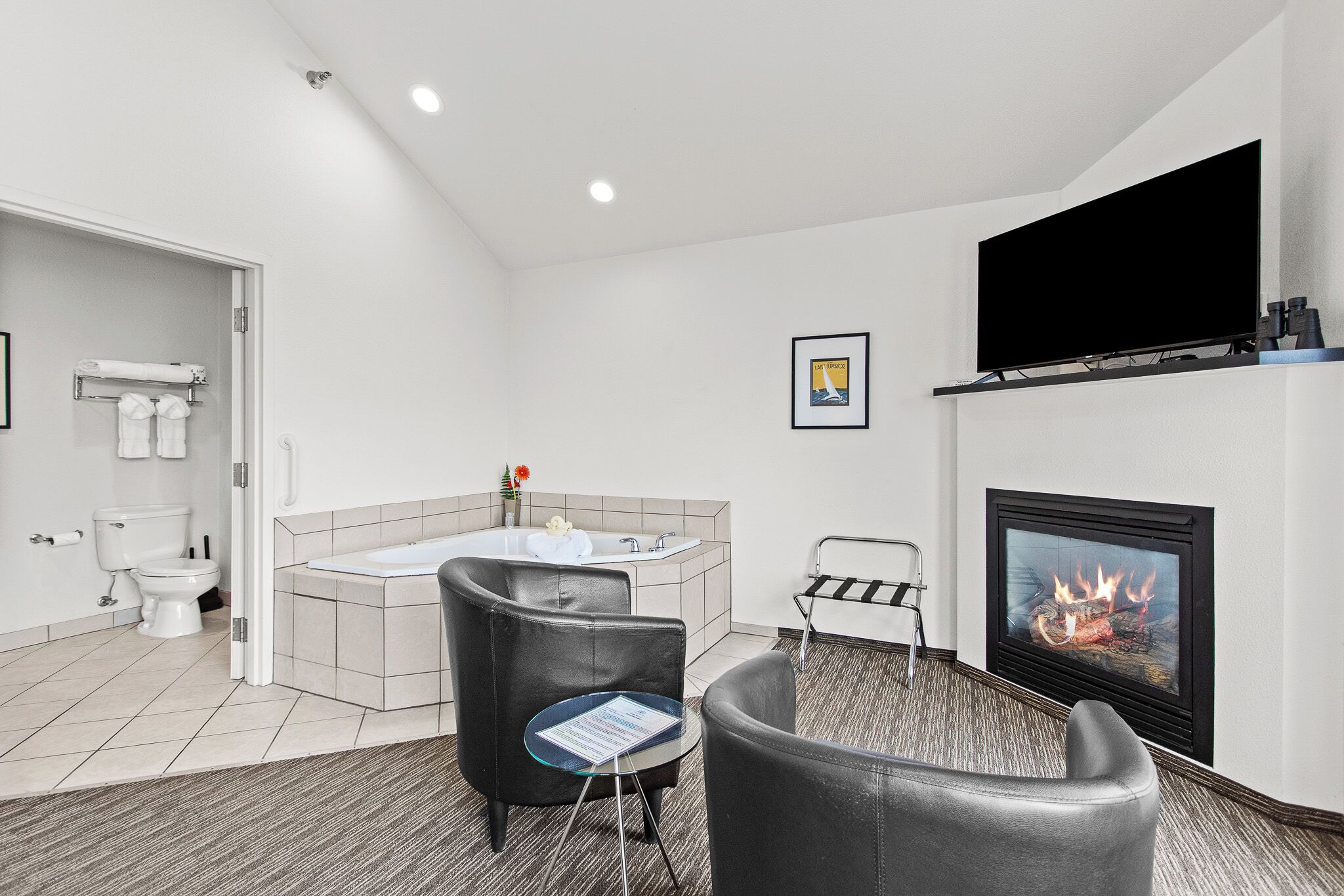 A modern bathroom with a toilet, sink, and bathtub. There is a fireplace with a fire, a television mounted on the wall, and a small table with a chair.