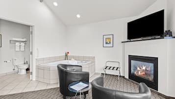 A modern bathroom with a toilet, sink, and bathtub. There is a fireplace with a fire, a television mounted on the wall, and a small table with a chair.