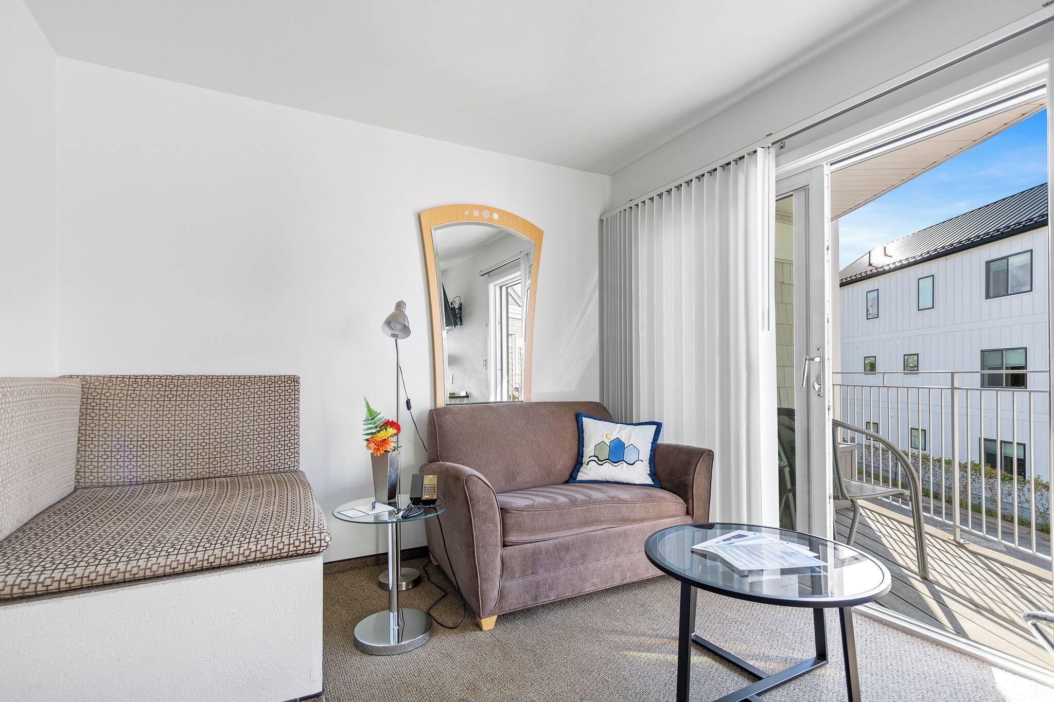 Una habitación de hotel moderna con sofá, una mesita, un espejo y un balcón con vistas a un edificio.