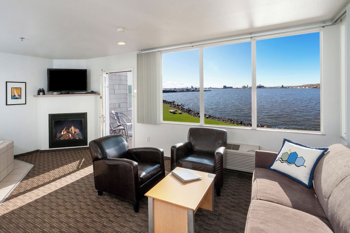 A modern living room with a fireplace, leather chairs, a sofa, and a large window with a view of the water.