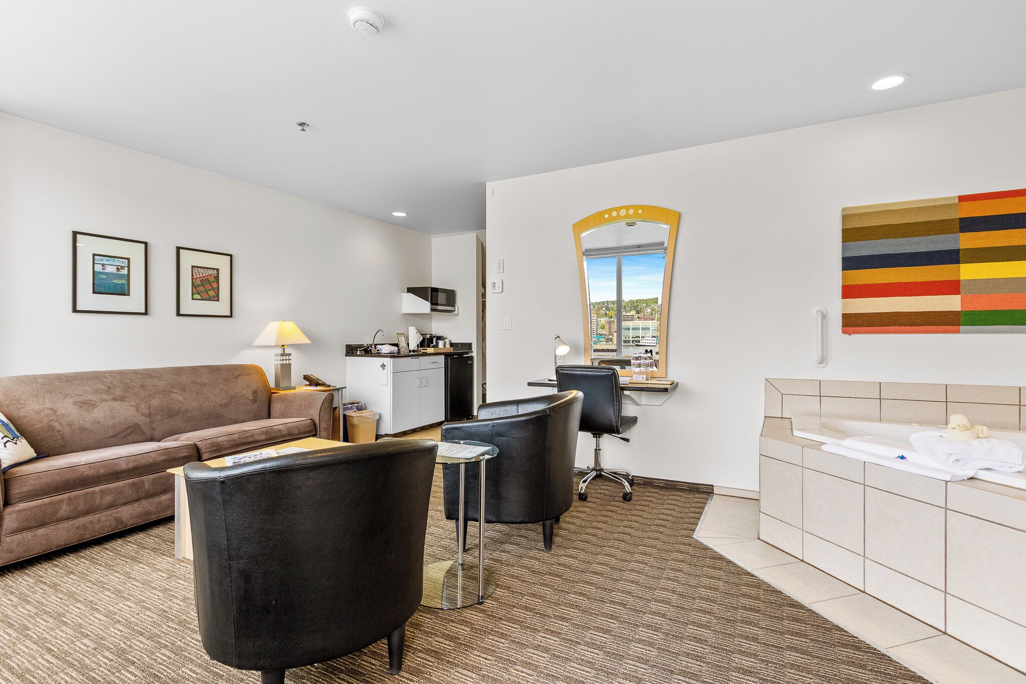 Habitación de hotel con sofá, dos sillones, escritorio, espejo, baño con bañera y vista a la ciudad.