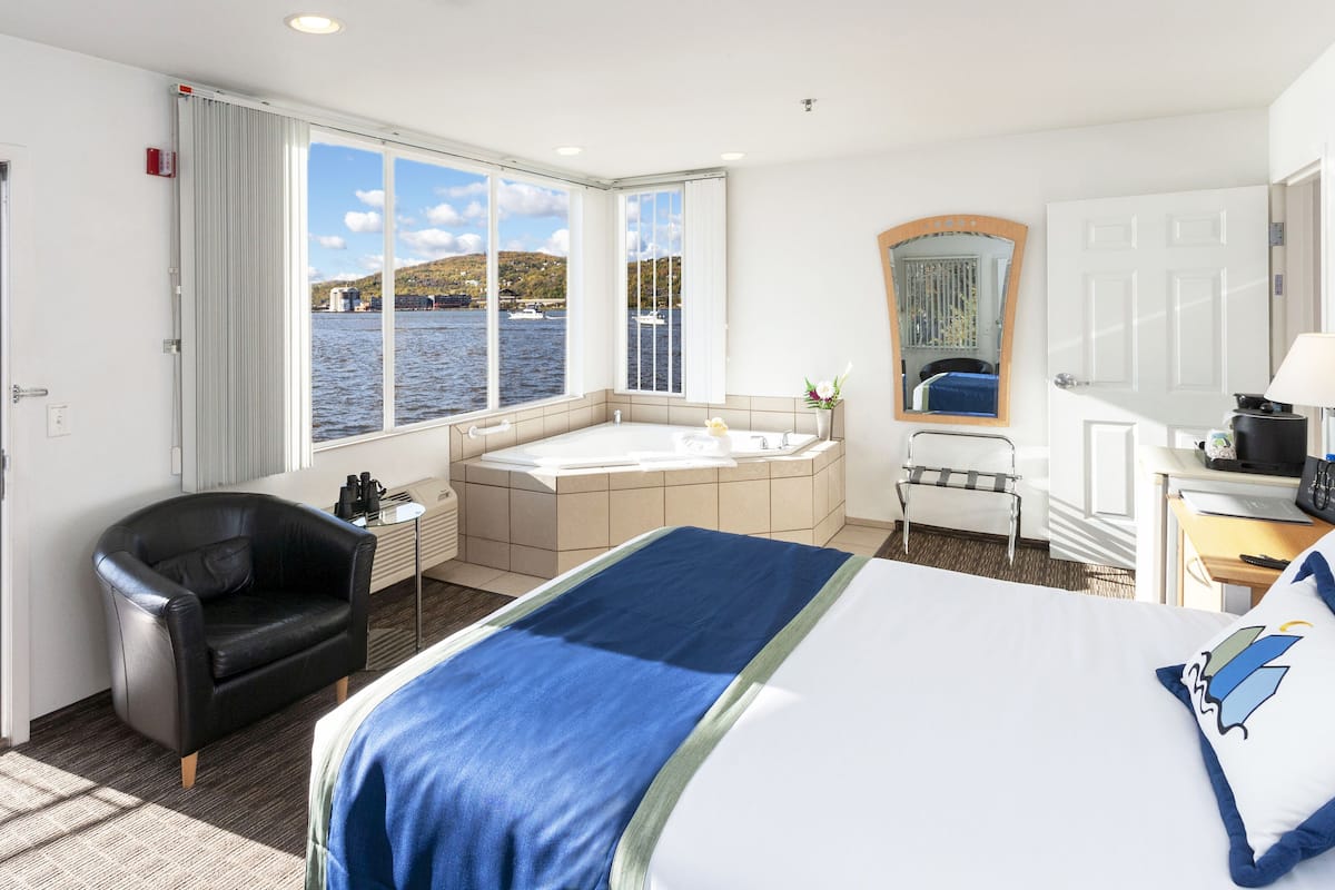 A hotel room with a bed, a desk, a chair, a bathtub, and a view of the water.