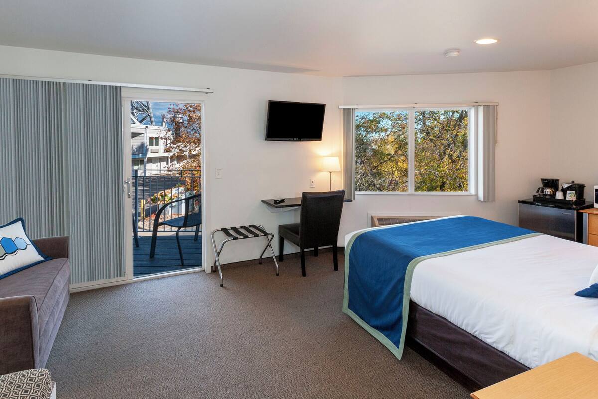 A hotel room with a bed, a desk, a chair, a TV, and a balcony with a view of buildings and trees.