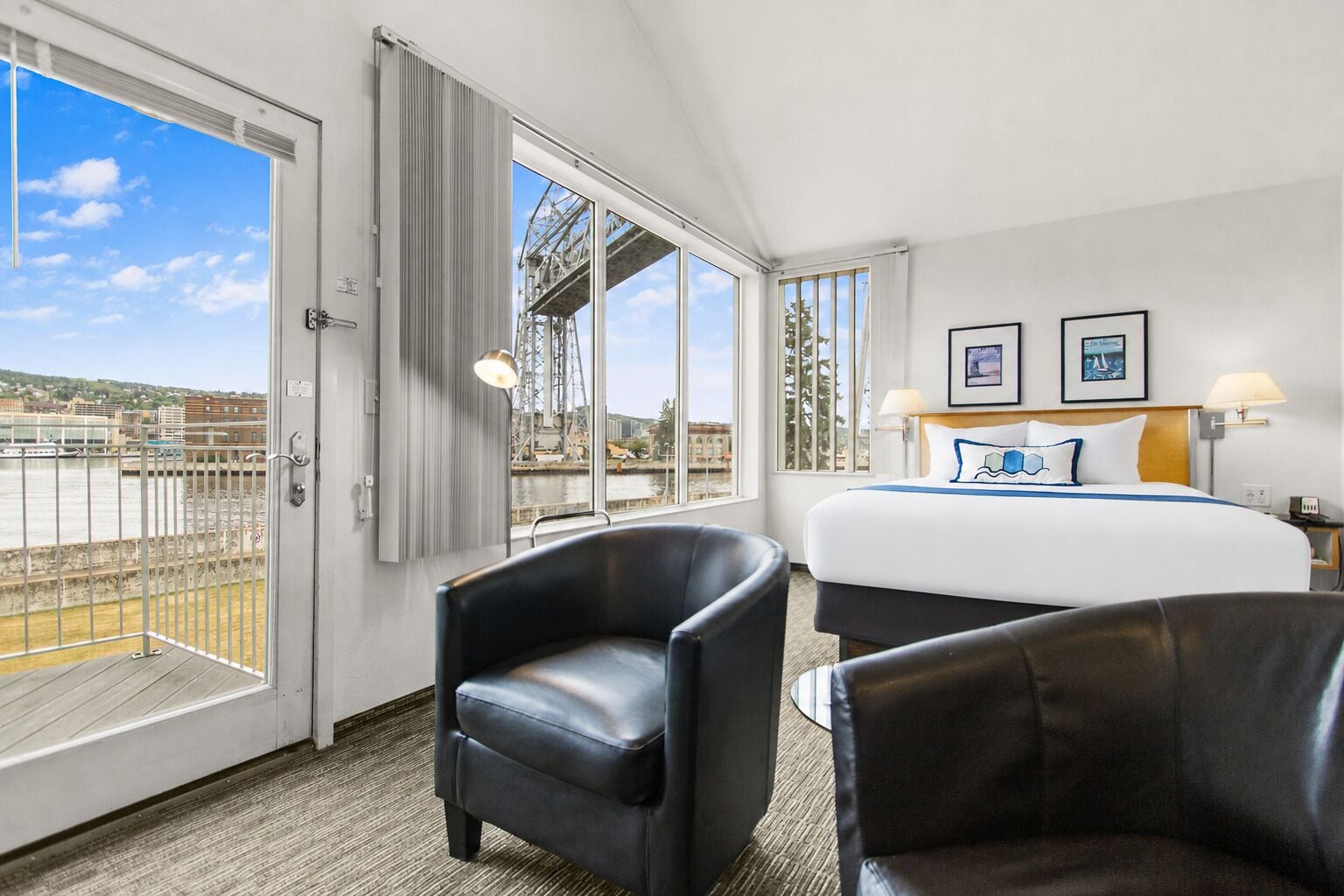 Quarto de hotel com uma cama, duas cadeiras, uma escrivaninha, uma luminária e uma janela grande com vista para uma ponte e para a água.