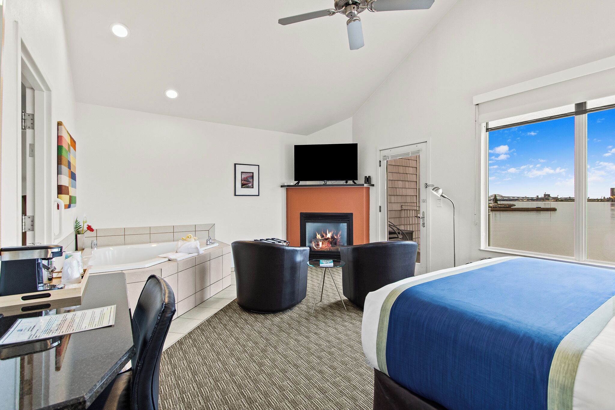 Habitación de hotel con cama, escritorio con computadora, silla, chimenea y ventana grande con vistas.