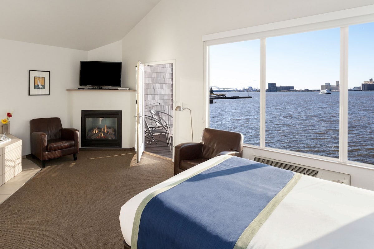 A hotel room with a large bed, a fireplace, a TV, and a view of the water.