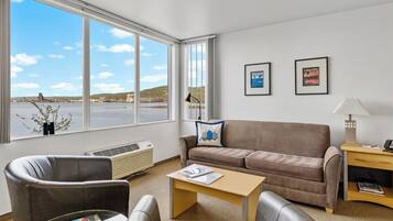Un salon comprenant un canapé, deux fauteuils, une table basse, une lampe et un radiateur. Deux tableaux encadrés ornent le mur, et une fenêtre offre une vue sur un plan d’eau et des collines.