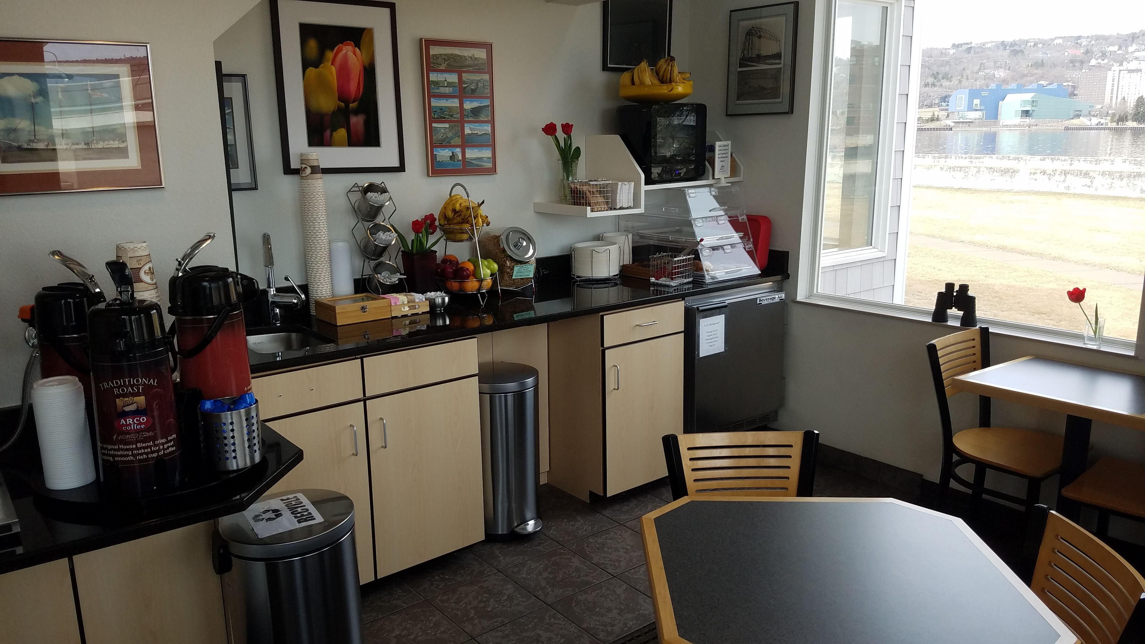 A kitchen with a coffee machine, a trash bin, and a window with a view of buildings.
