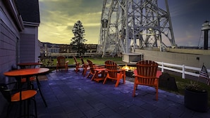 Un patio exterior con sillas Adirondack y un pozo de fuego, con vistas a una gran estructura metálica tipo puente.