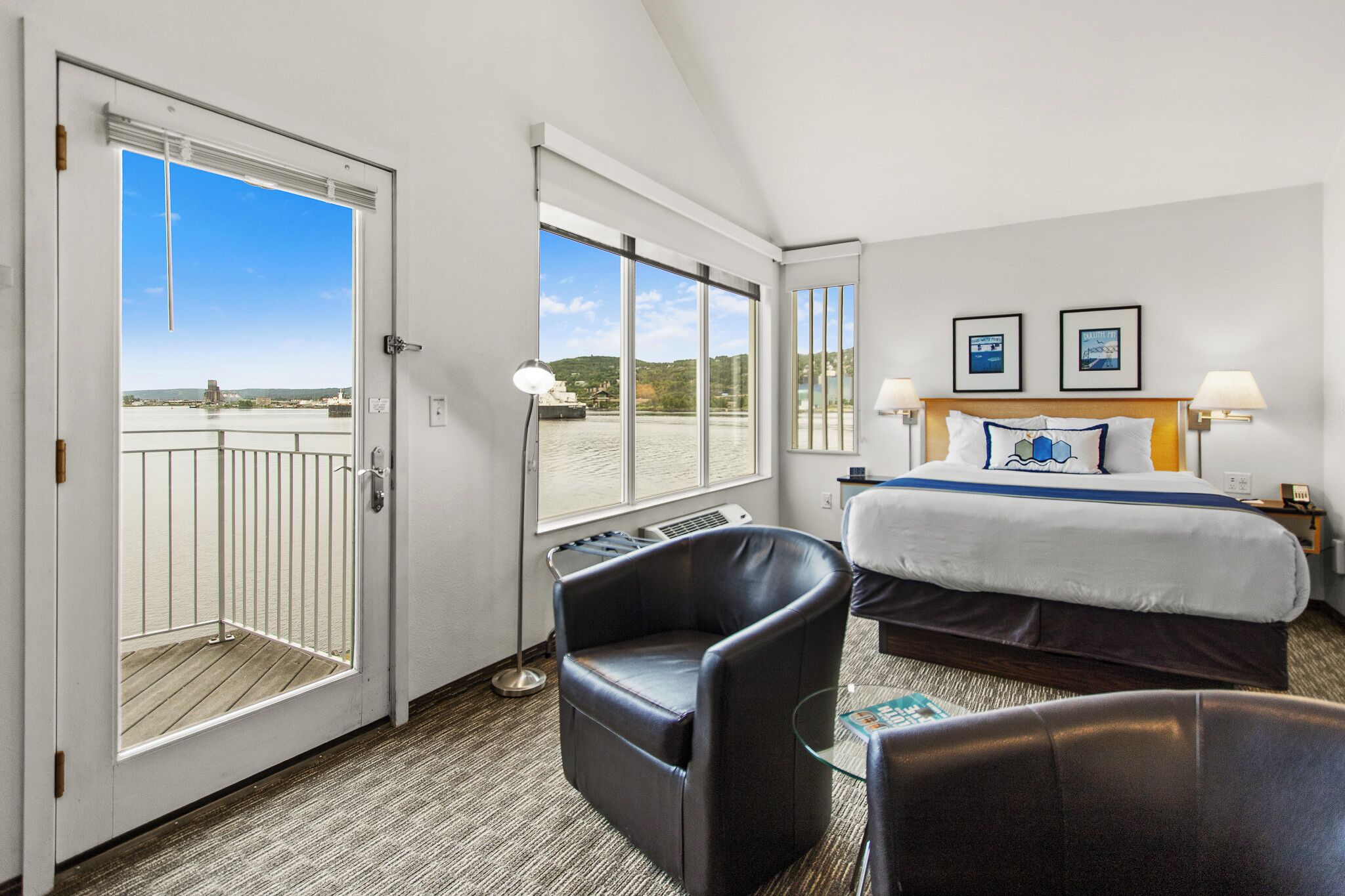 Habitación de hotel con cama, dos sillas, escritorio, ventana con vista al mar y puerta que da a un balcón.