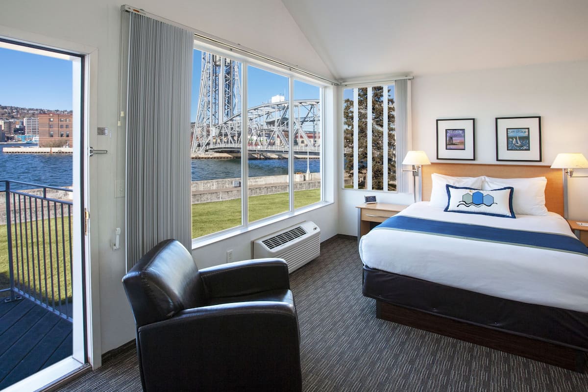 A hotel room with a large bed, a desk, a chair, and a view of a water body and industrial structures.