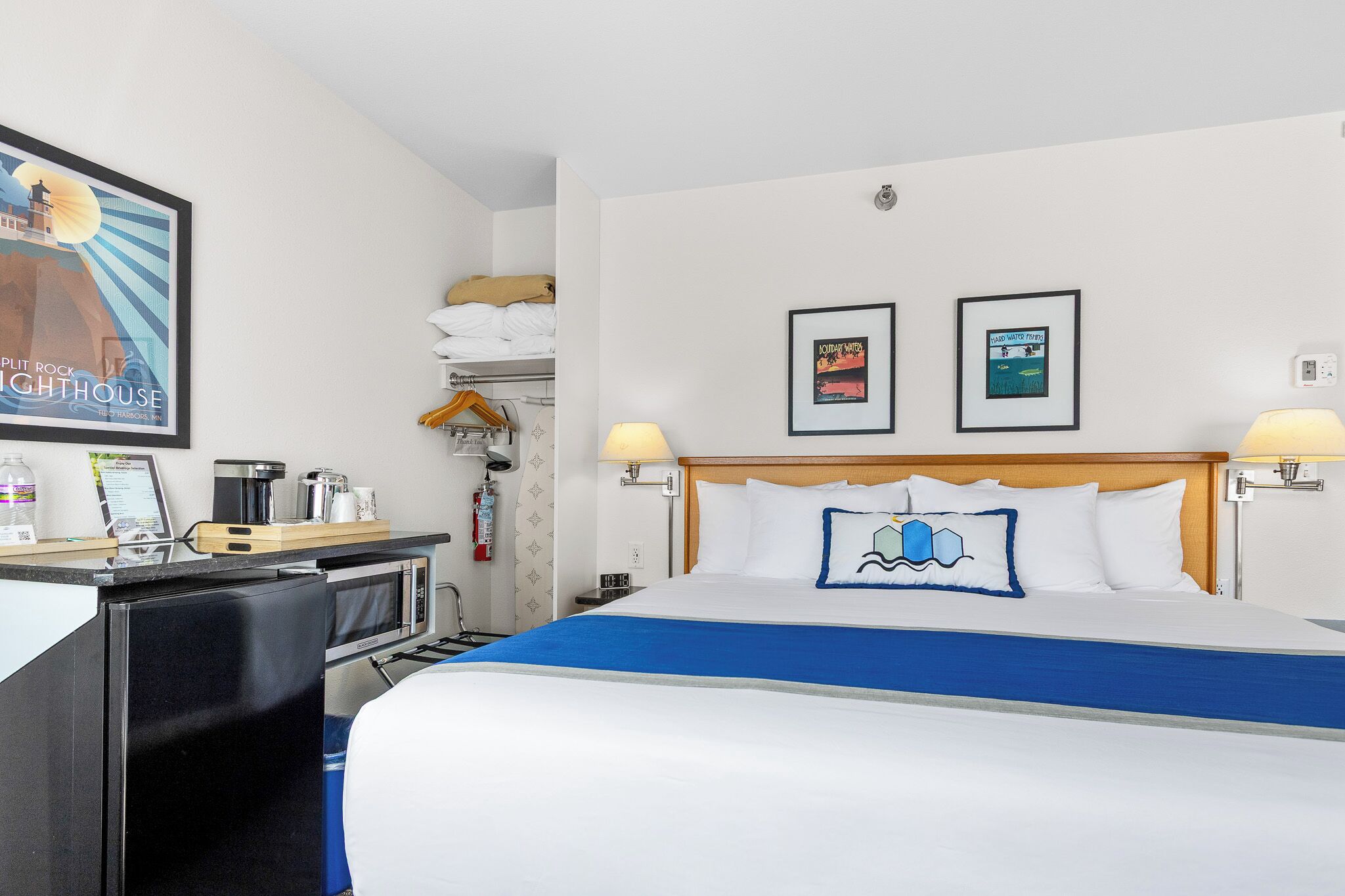 A hotel room with a bed, bedside lamps, a desk with a coffee maker, a microwave, and a framed picture of a lighthouse.