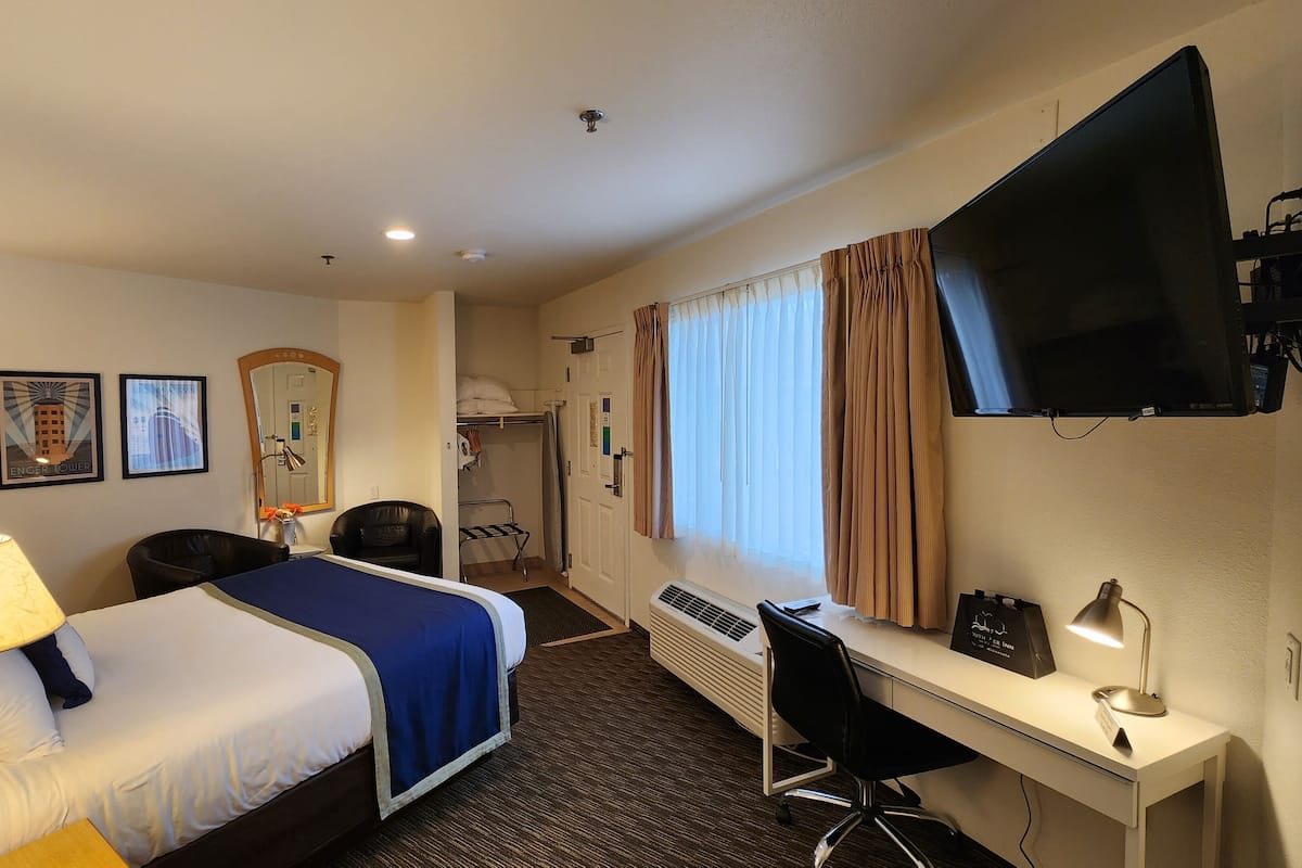 A hotel room with a bed, desk, chair, TV, and artwork on the wall.