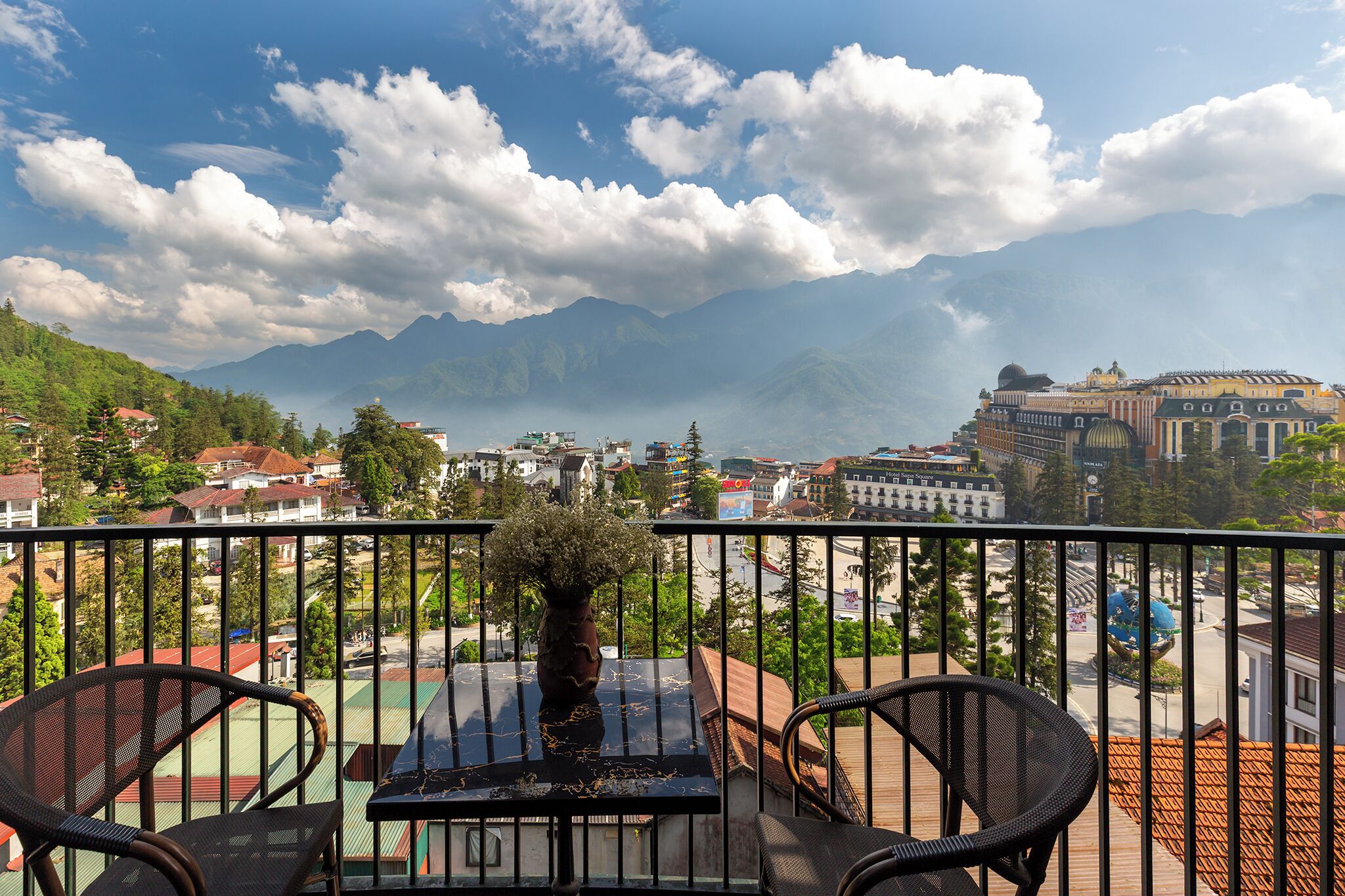 A balcony with chairs and a table, overlooking a cityscape with mountains in the background.