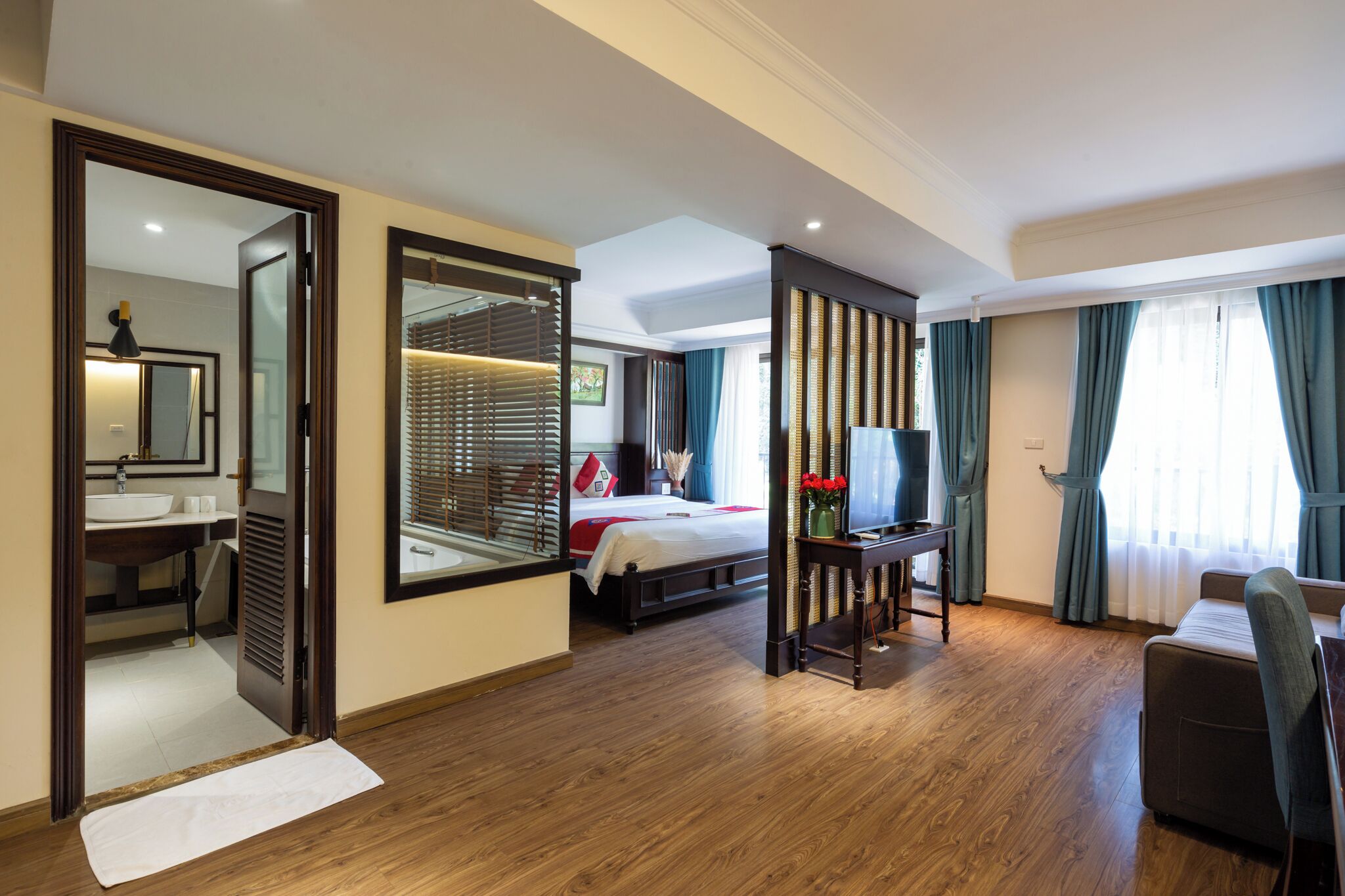 Quarto de hotel com cama, escrivaninha, televisão, banheiro com espelho e uma janela grande com cortinas.