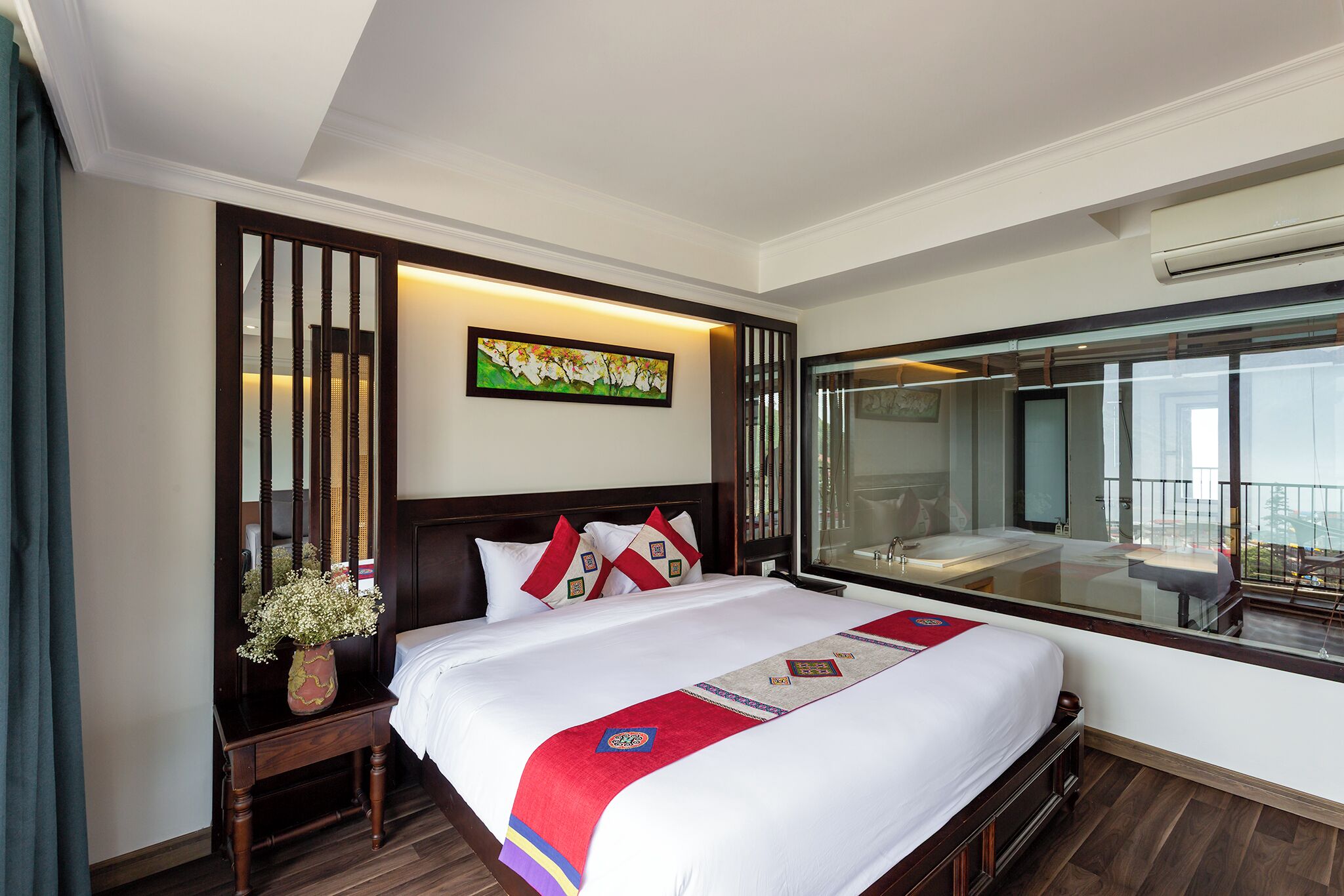A hotel room with a large bed, a bedside table with a vase, a painting on the wall, and a large window with a view.