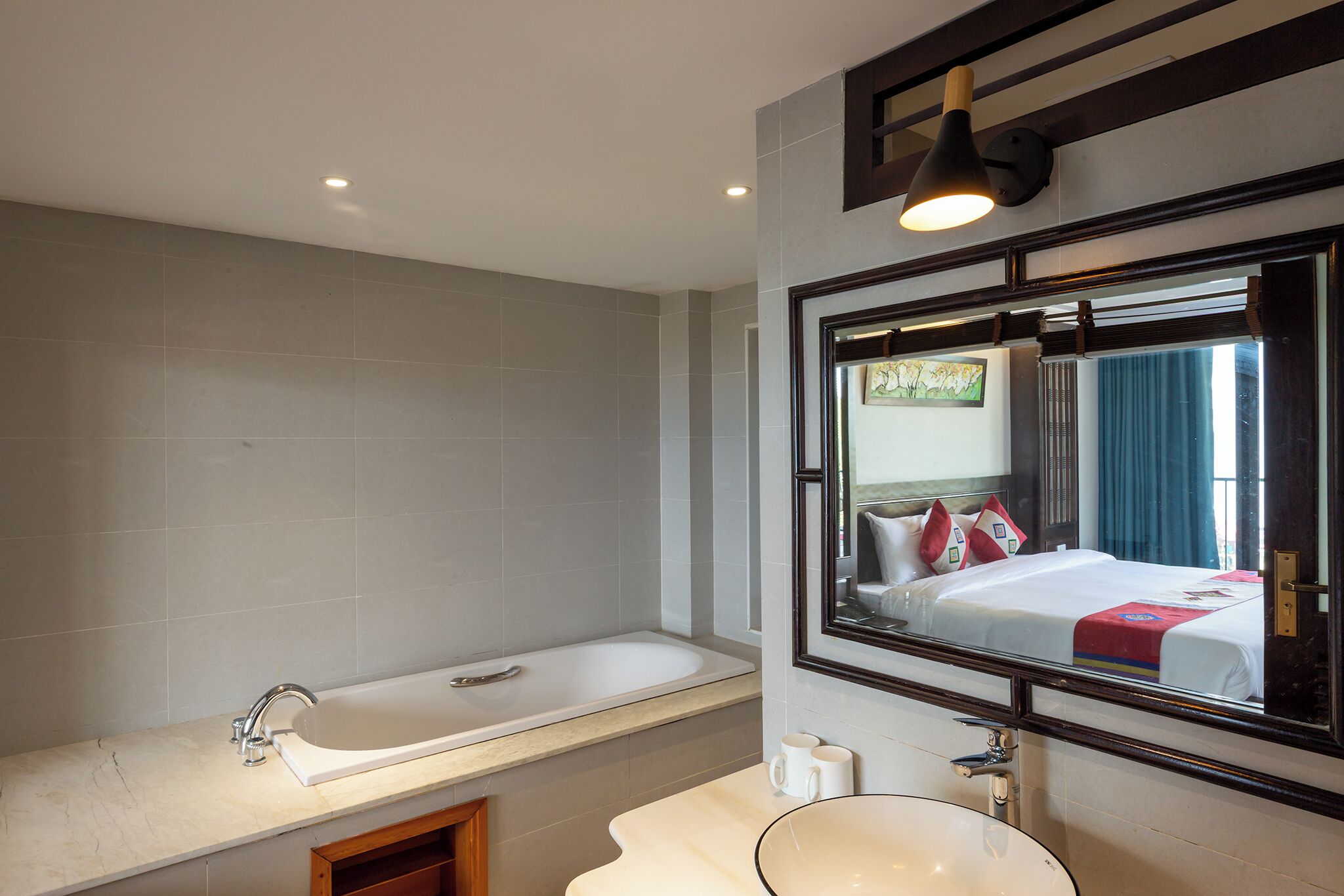 A bathroom with a bathtub, a sink, and a large mirror reflecting a bed with pillows.