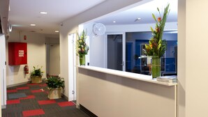 A hallway with a red and black carpet, a reception desk with potted plants, and a fire extinguisher on the wall. - Sydney Student Living - Hostel (Concord)