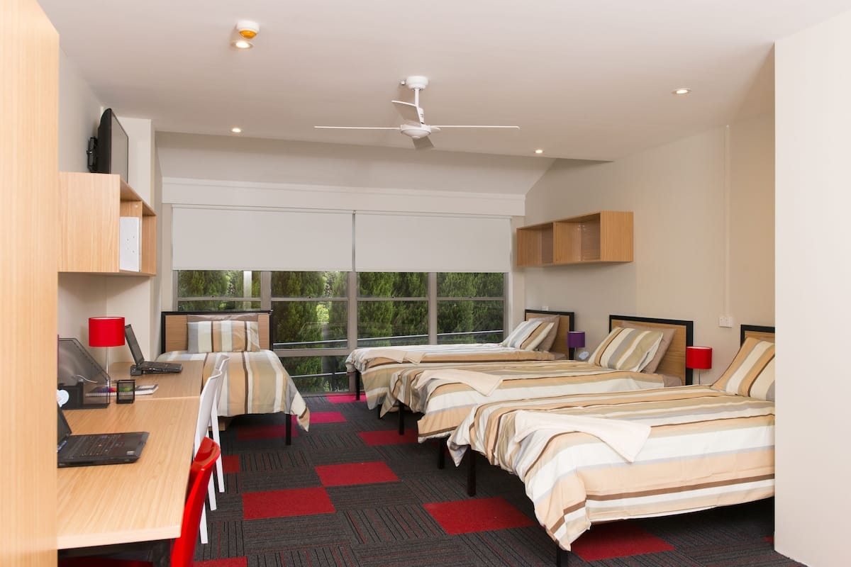 Habitación de hotel con dos camas individuales, un escritorio con una computadora portátil, una silla, una lámpara roja y un ventilador de techo.