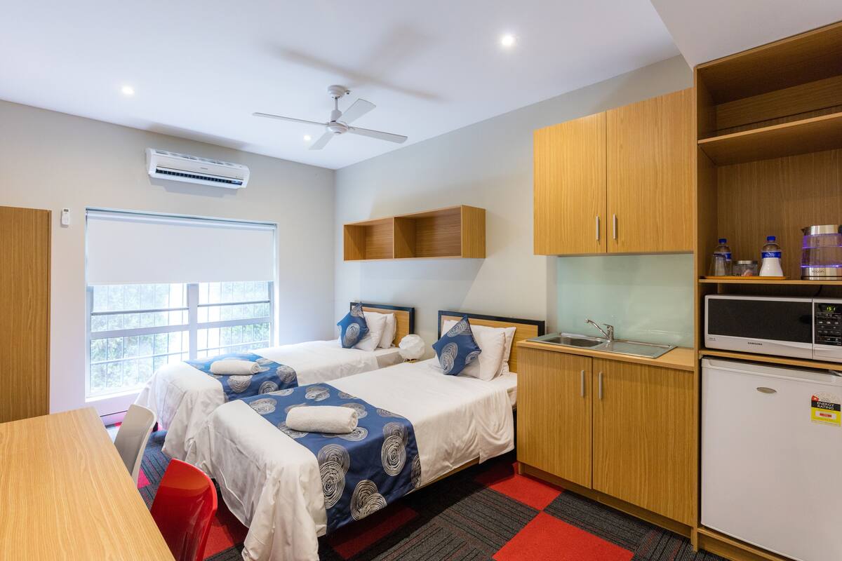Habitación de hotel con dos camas, kitchenette, ventilador de techo y ventana con vistas.