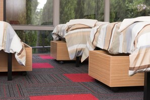 A hotel room with four beds arranged in a row, each with a striped blanket and a wooden headboard. The room has a carpeted floor with red and gray patterns, and large windows offering a view of trees outside.