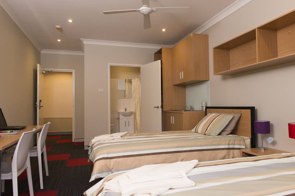 Habitación de hotel con dos camas, un escritorio, una silla, un ventilador de techo y un baño visible a través de una puerta abierta.