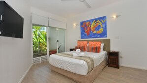 A modern bedroom with a large bed, a ceiling fan, a TV mounted on the wall, and a window with a view of greenery. - Seascape Holidays- Beaches (Port Douglas)