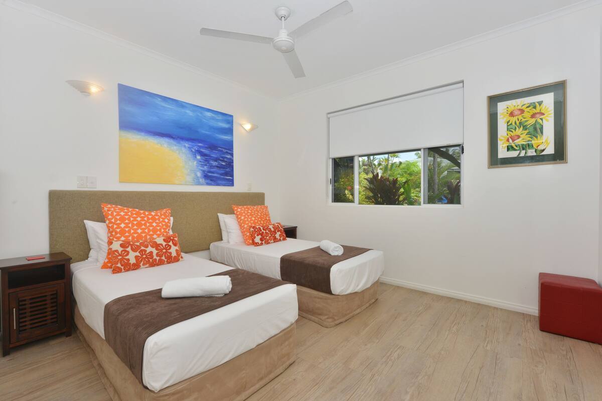 A hotel room with two beds, a ceiling fan, a painting of a beach, a window with a view of greenery, and a framed picture of sunflowers.
