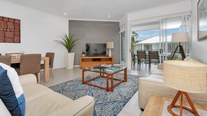 A modern living room with a sofa, coffee table, TV, and dining area. - Seascape Holidays- Beaches (Port Douglas)