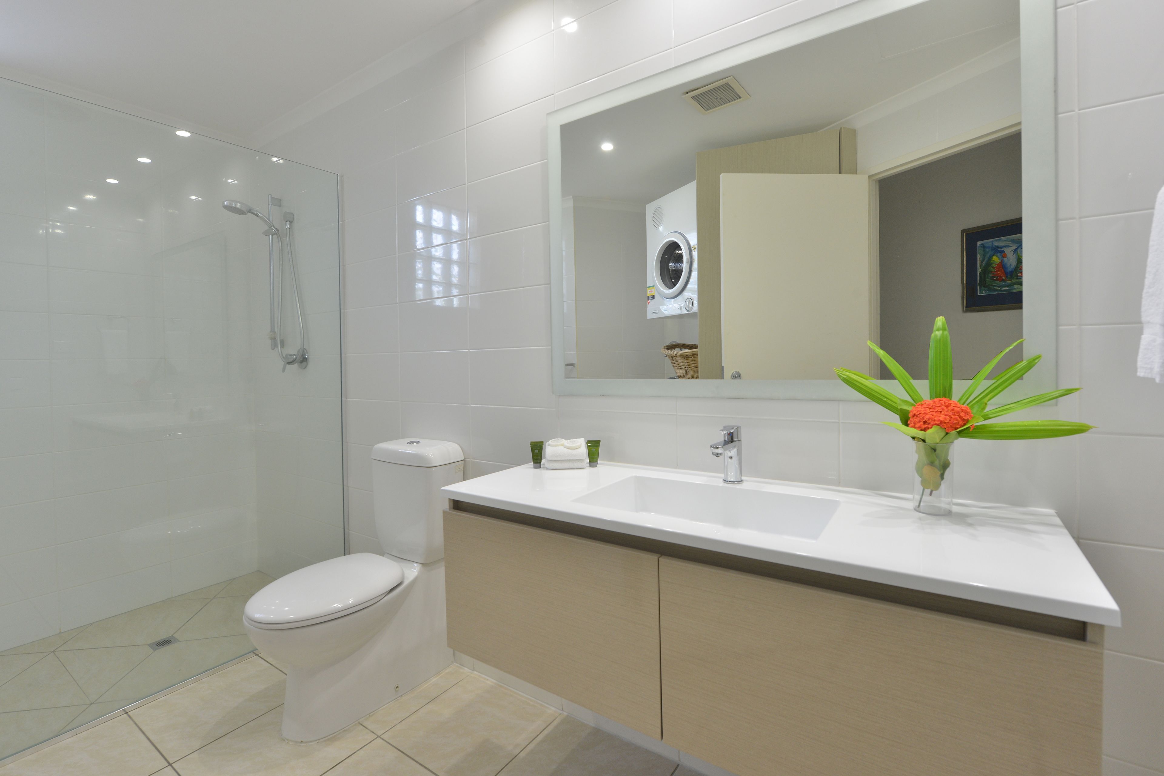 Een moderne badkamer met een douchecabine van glas, een toilet, een wastafel met spiegel en een vaas met bloemen op het aanrecht.
