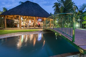 A pool with a bridge, a thatched-roof building, and lush greenery. - The Royal Villas Swaziland (Lobamba)