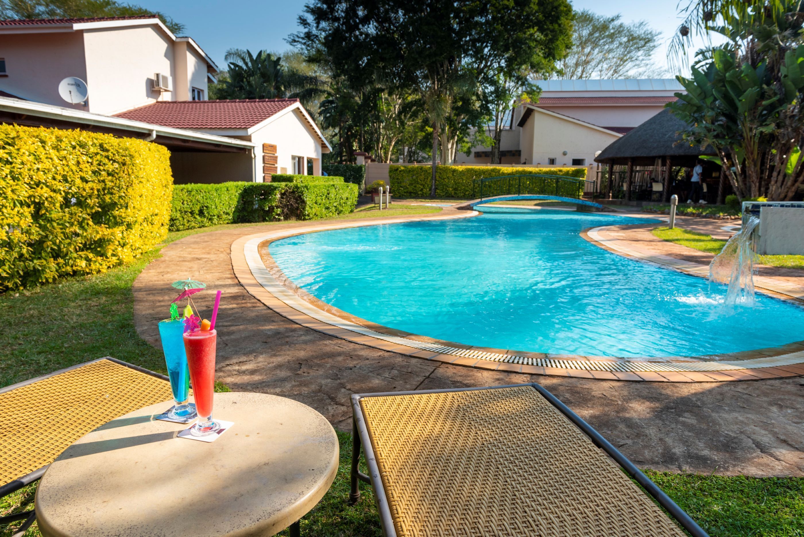 Una piscina con sillones de descanso y una mesa, rodeada de setos cuidados y un edificio con tejado rojo.