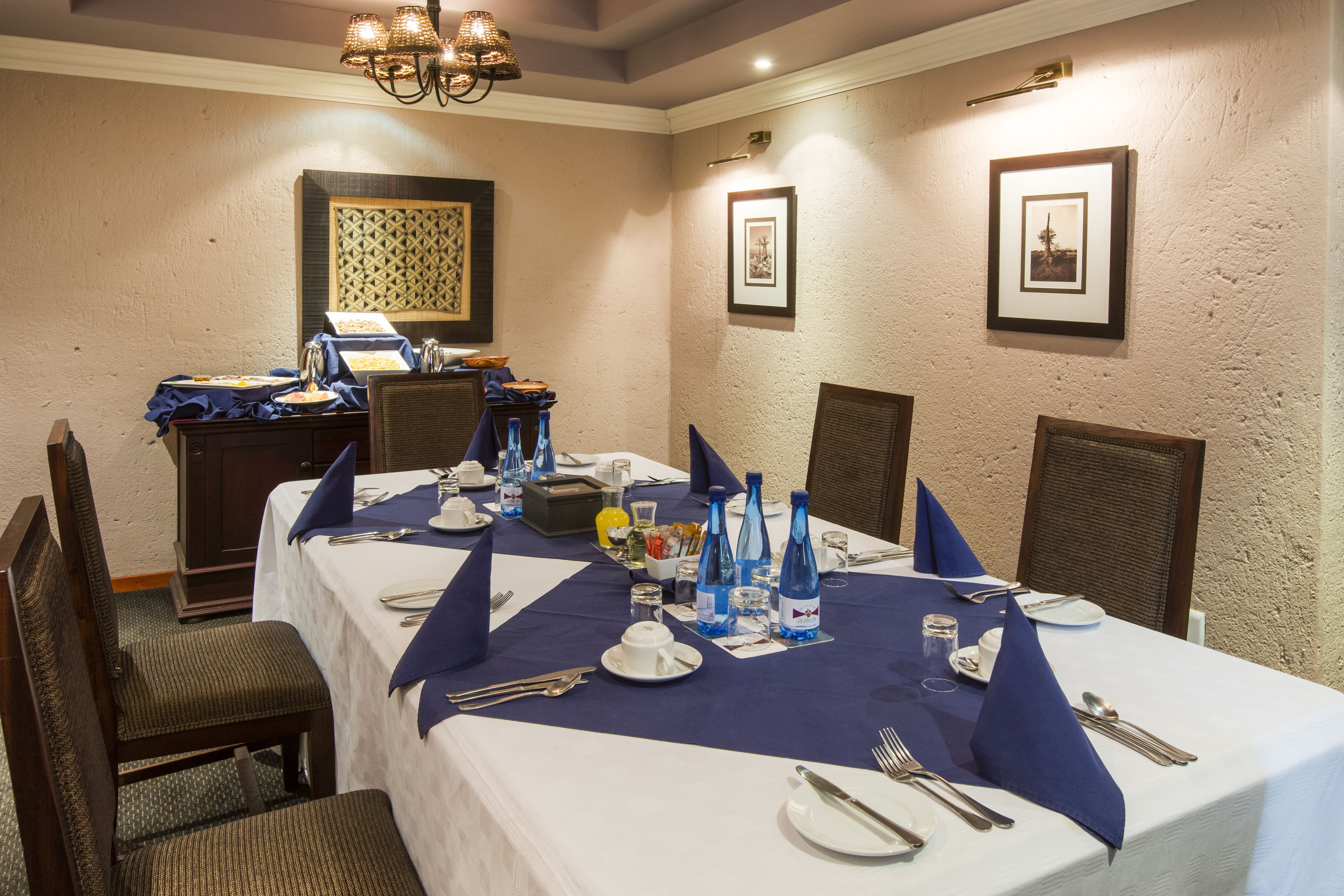 A dining area with a long table set for a meal, chairs, a chandelier, framed pictures on the walls, and a buffet station in the background.